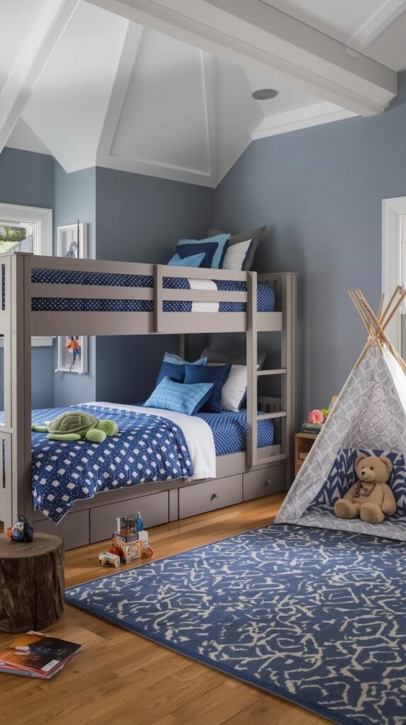 Charming boys’ bedroom with an adventurous, cozy feel. The walls are painted a soft slate blue with white trim and angled ceilings, creating a whimsical nook-like effect. A gray wooden bunk bed with blue polka-dot bedding and plush pillows anchors the room, featuring a stuffed turtle toy and accent cushions in navy and sky blue. To the side, a small indoor teepee tent with geometric patterns and a plush teddy bear inside adds a playful touch. The space includes a patterned blue and cream area rug, a wooden stump side table, and scattered children’s toys and books for a lived-in, imaginative vibe. Natural light streams through sheer curtains, enhancing the room’s serene, storybook atmosphere.