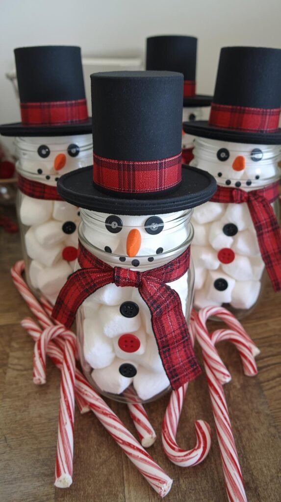 Snowman mason jars filled with marshmallows, each decorated with buttons, plaid ribbon scarves, and black paper hats. Arranged on a countertop beside candy canes