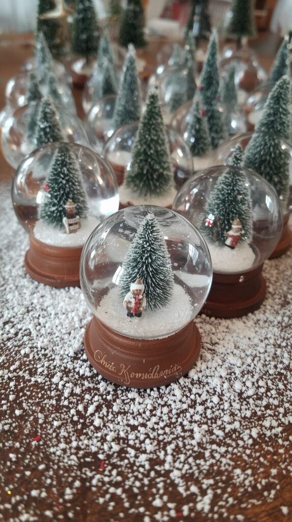 Mini snow globes with glitter, bottle brush trees, and figurines sitting on a craft table sprinkled with faux snow.