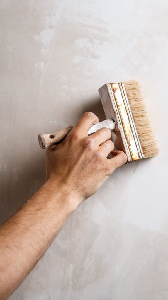 Close-up shot of a hand applying lime wash paint to a wall using a wide natural bristle brush, soft neutral taupe limewash strokes visible across the surface, overlapping cloudlike patterns, matte texture, subtle lighting highlighting brush movement, realistic skin tones.