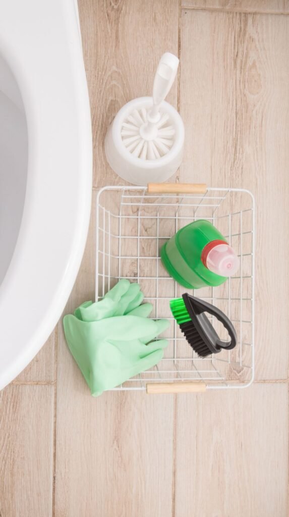 Flat-lay of bathroom cleaning essentials arranged neatly: toilet bowl cleaner, scrub brush, disinfecting wipes, gloves, microfiber cloths.