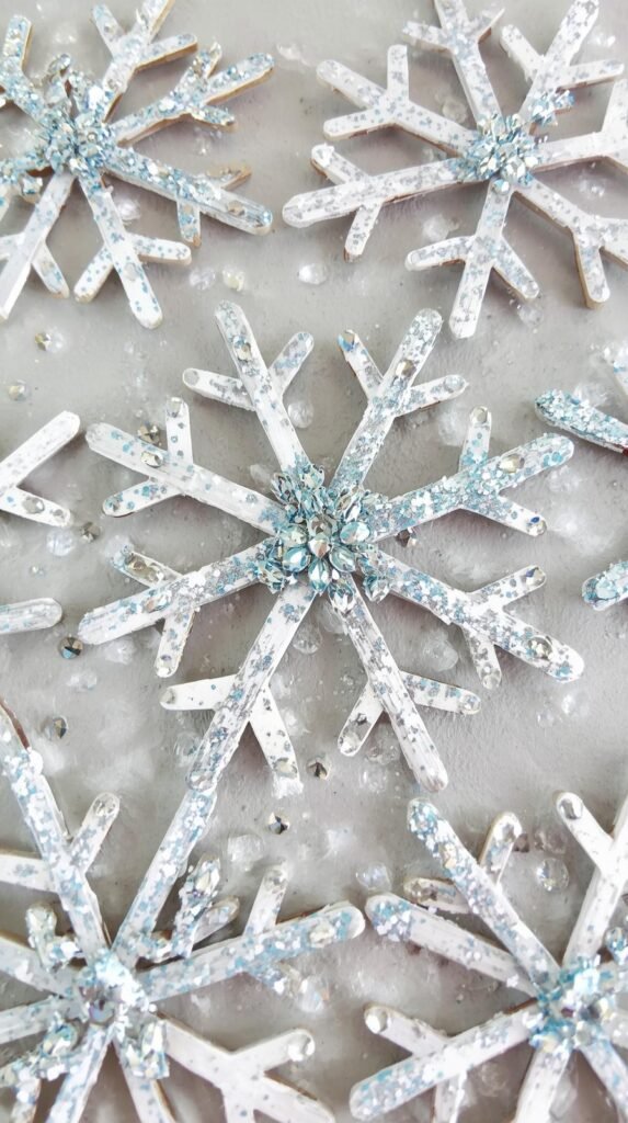 Wooden craft sticks arranged into snowflake shapes, painted white and blue with added glitter and adhesive gems.