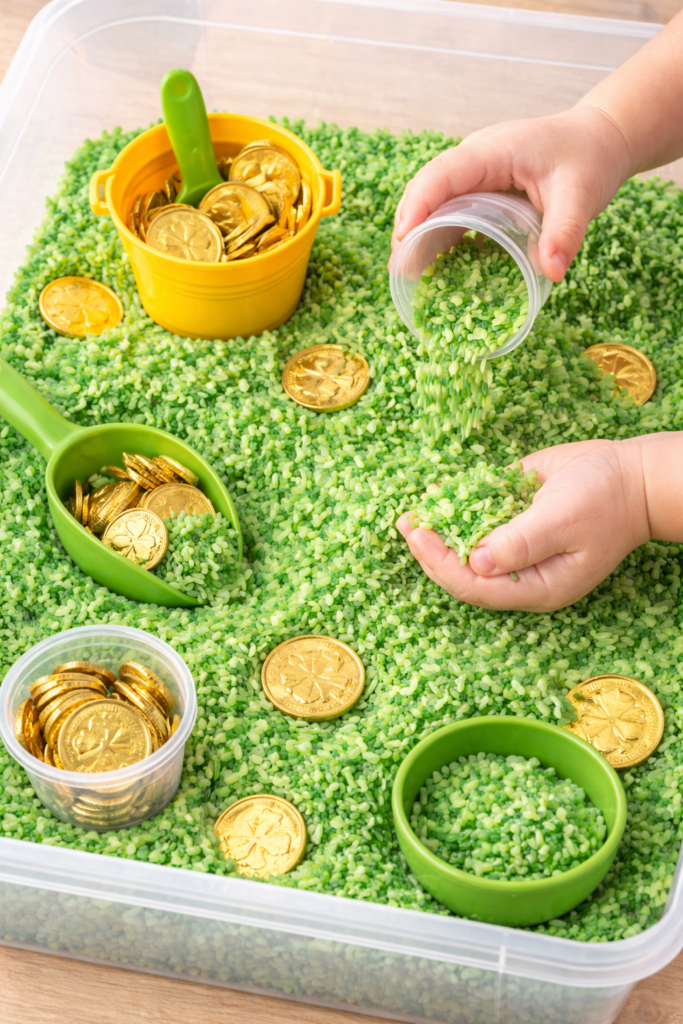 clear plastic bin filled with green dyed rice, gold coins, small scoops and cups